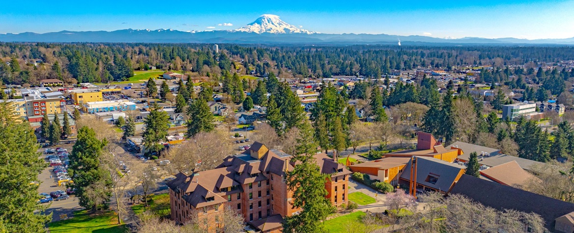 An aerial view of PLU's upper campus, with the red brick Harstad Hall and Anderson University Center at the bottom of the photo. At the top of the photo is Mount Rainier and blue sky.