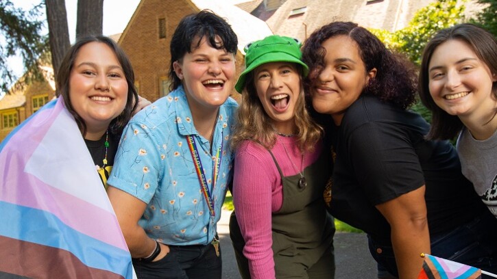 A group of 5 students stand together outside on the PLU campus, slightly crouched and smiling at the camera. One is holding a small pride flag and another has a large pride flag wrapped around their shoulders.