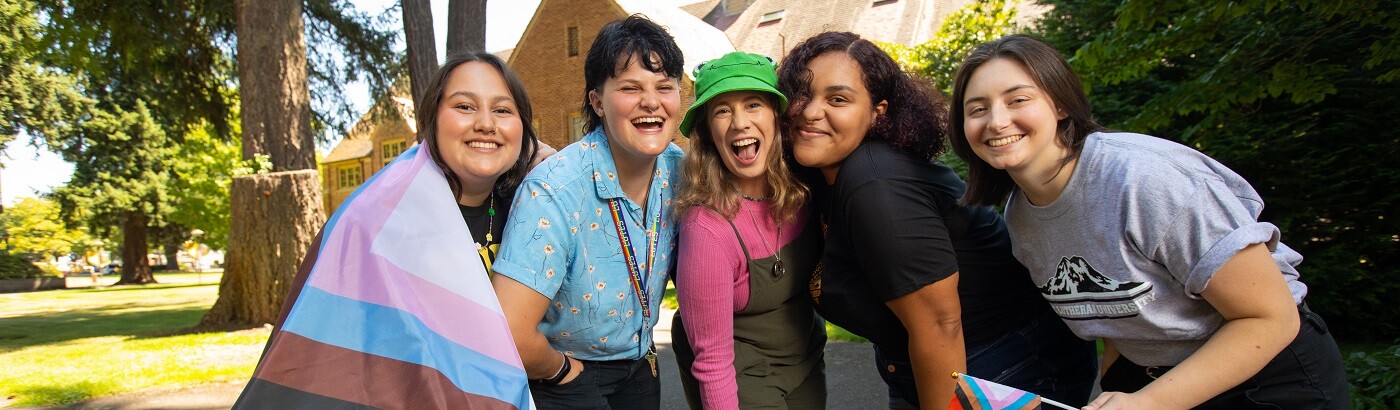 A group of 5 students stand together outside on the PLU campus, slightly crouched and smiling at the camera. One is holding a small pride flag and another has a large pride flag wrapped around their shoulders.