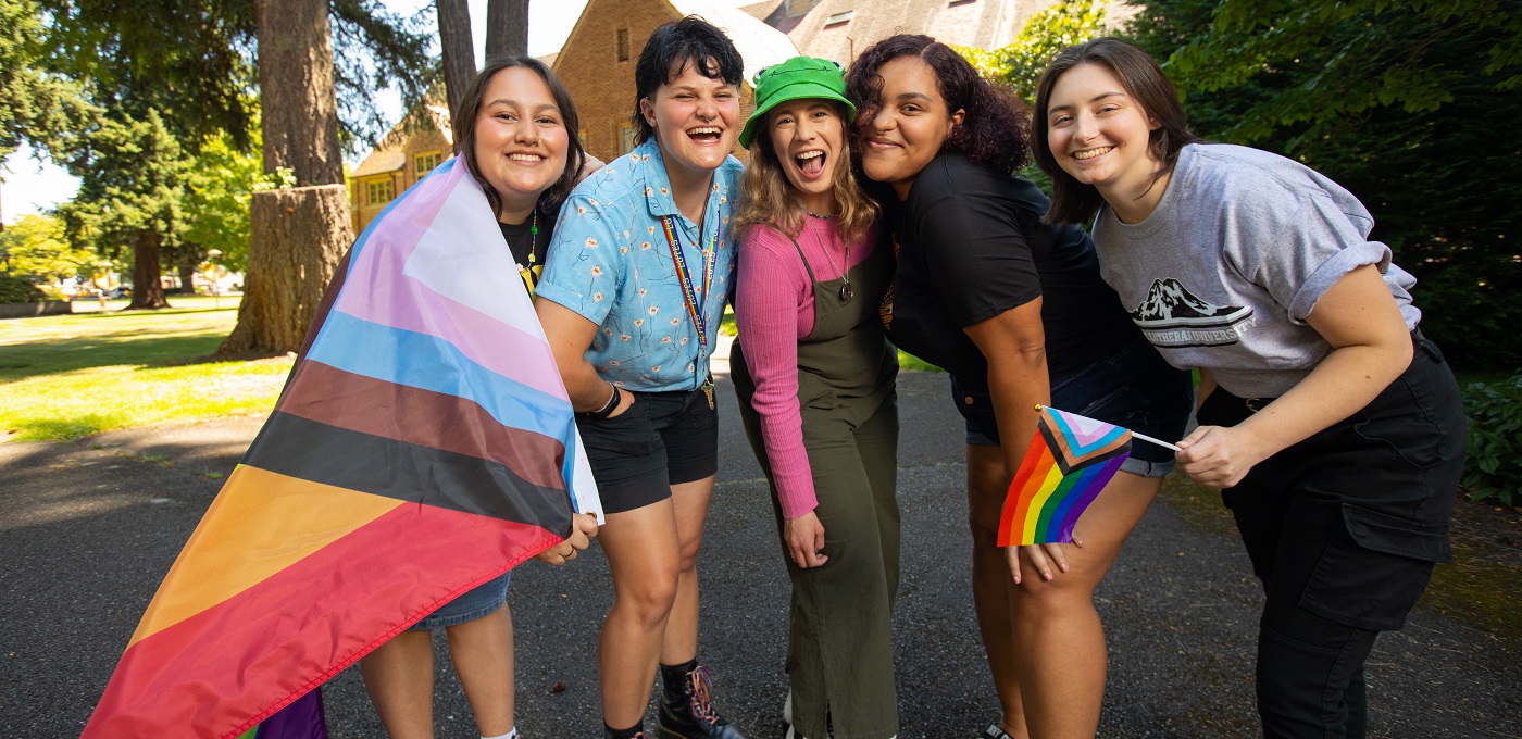 A group of 5 students stand together outside on the PLU campus, slightly crouched and smiling at the camera. One is holding a small pride flag and another has a large pride flag wrapped around their shoulders.