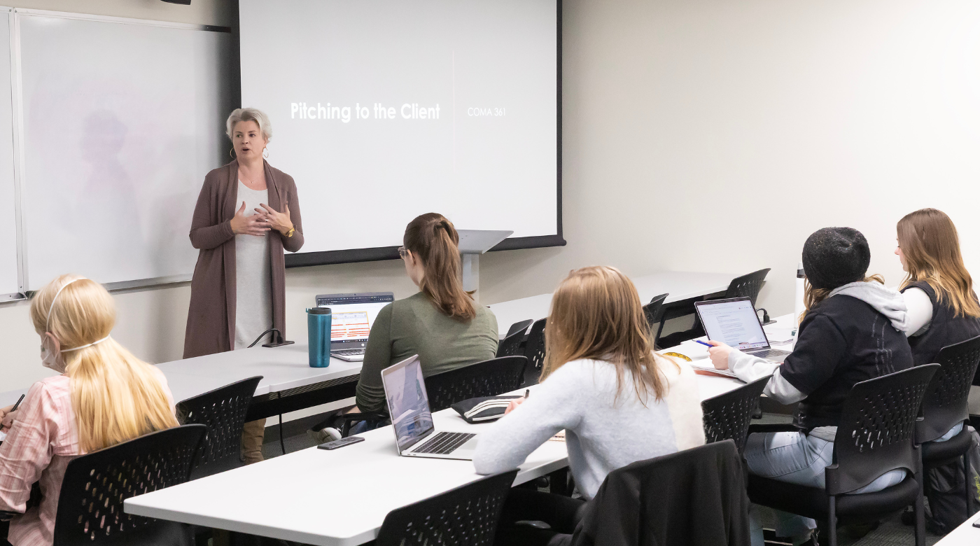 A professor stands in front of a classroom of students. There is a powerpoint presentation behind her that reads "How to pitch a client"