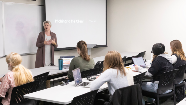 A professor stands in front of a classroom of students. There is a powerpoint presentation behind her that reads "How to pitch a client"