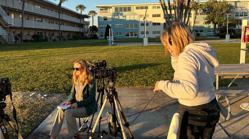Two women film outside in Florida. One is interviewing someone off camera and the other is listening with headphones.