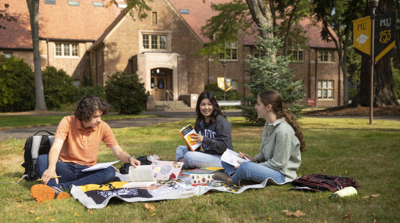 Three students sit on a blanket outside on the PLU campus.