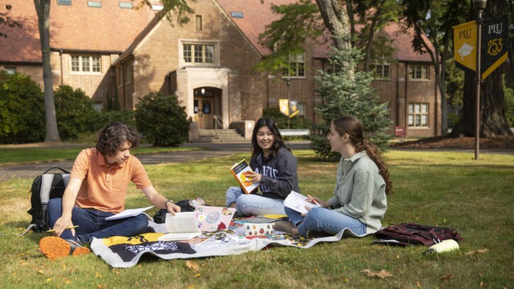 Three students sit on a blanket outside on the PLU campus.