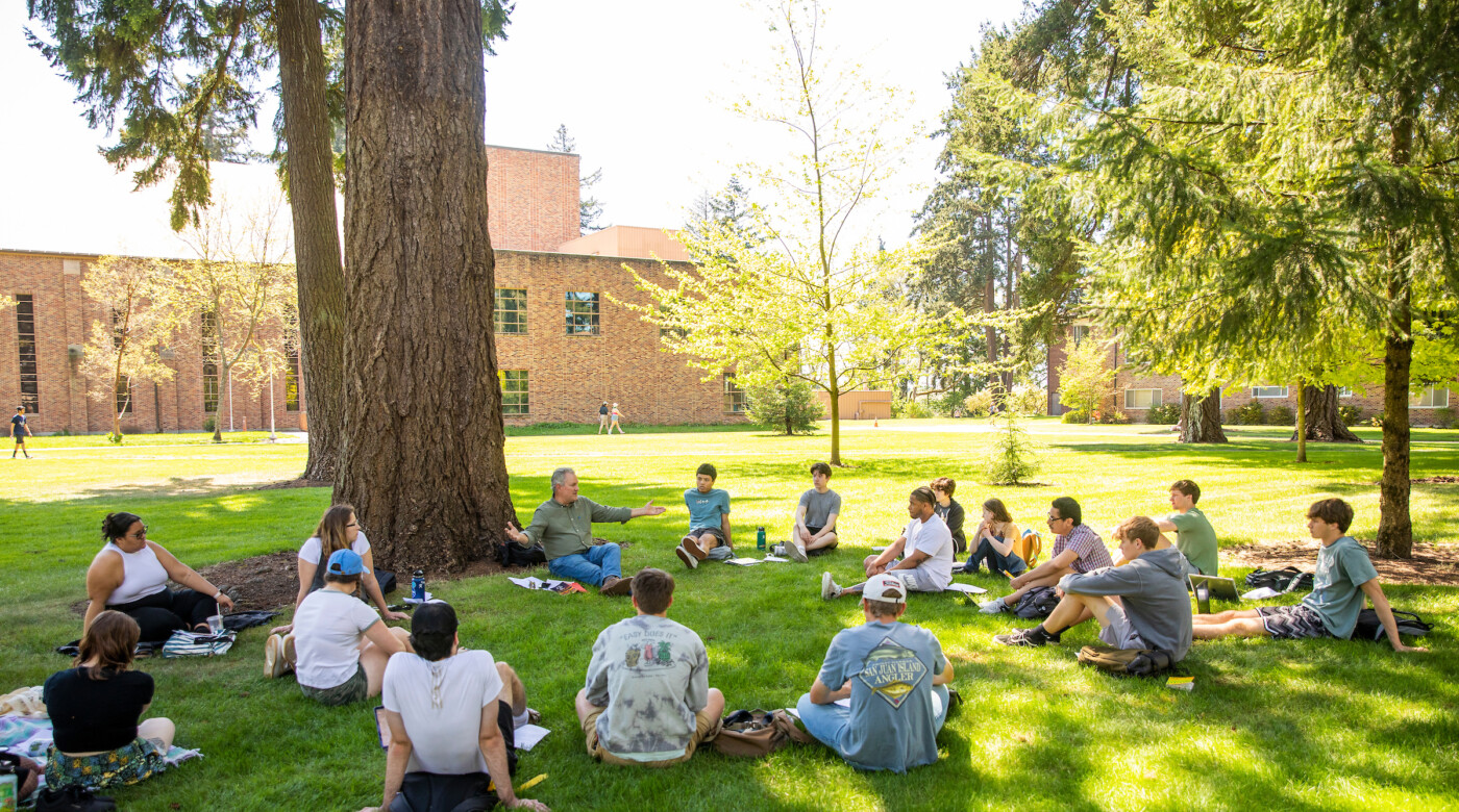 A professor conducts a class outside under trees, students sit in circle