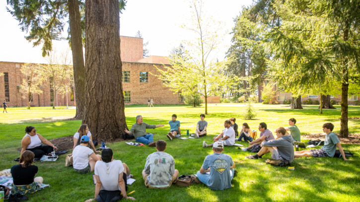 A professor conducts a class outside under trees, students sit in circle