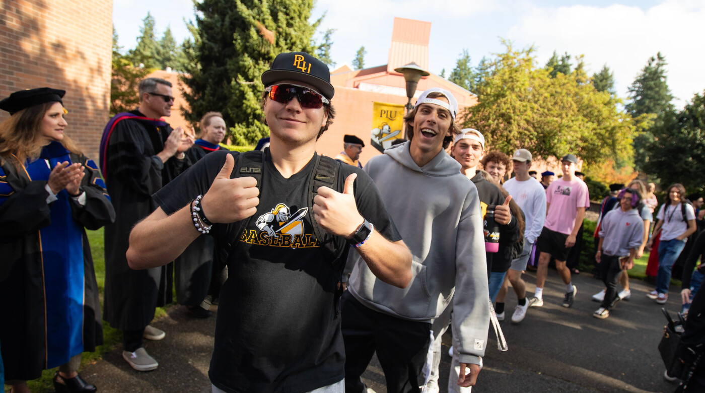 Students walking in convocation and one student smiles into camera and throws two thumbs up