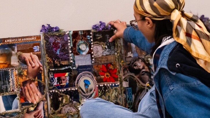 A student artist sits cross-legged on a floor in front of their artwork, which is a wide collage of sorts.