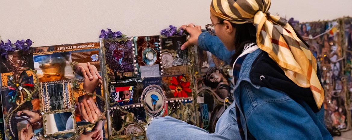 A student artist sits cross-legged on a floor in front of their artwork, which is a wide collage of sorts.