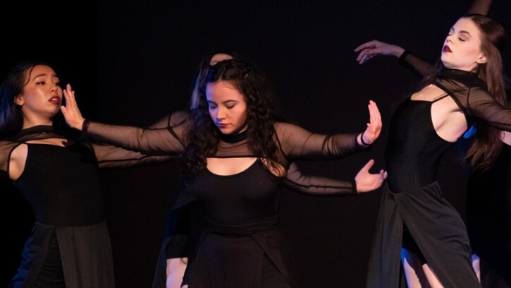 A group of dancers dressed in black camisoles, sheer black shoulder and arm tops and long black skirts in the middle of a dance performance. One dancer in the middle holds her arms out to each side, as if pushing the two dancers beside her away.