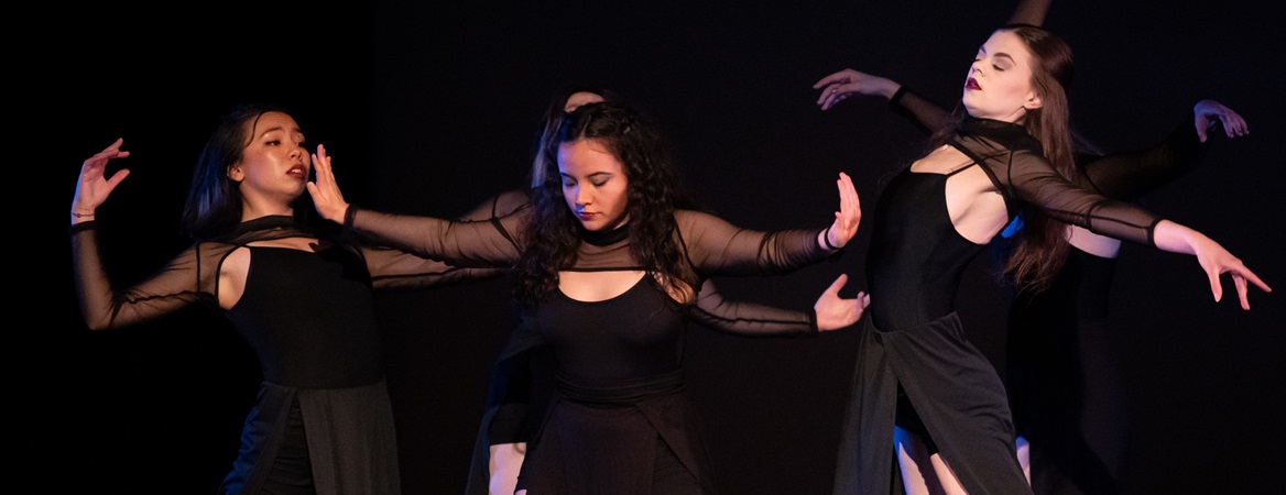 A group of dancers dressed in black camisoles, sheer black shoulder and arm tops and long black skirts in the middle of a dance performance. One dancer in the middle holds her arms out to each side, as if pushing the two dancers beside her away.