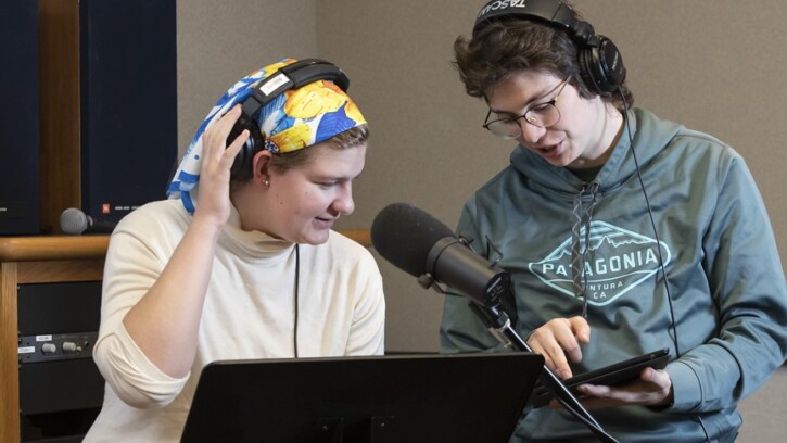 Two students collaborate in a recording studio on campus. Both are wearing headphones and one is sitting in front of a large microphone. The other student is holding a tablet, which both students are looking at.