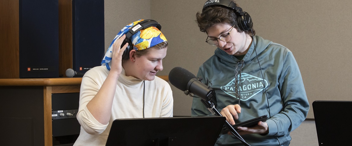 Two students collaborate in a recording studio on campus. Both are wearing headphones and one is sitting in front of a large microphone. The other student is holding a tablet, which both students are looking at.