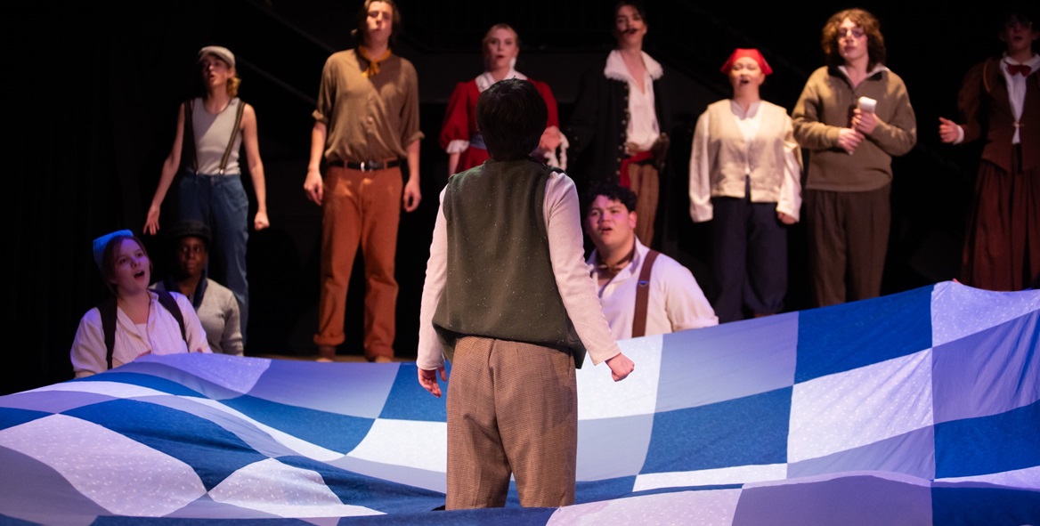 An image from the production of Peter and the Starcatcher. An actor stands in the middle of the stage, back to the audience, surrounded by a blanket of blue and white squares to represent water. Facing him are 10 other actors, some standing some kneeling, in the middle of singing.