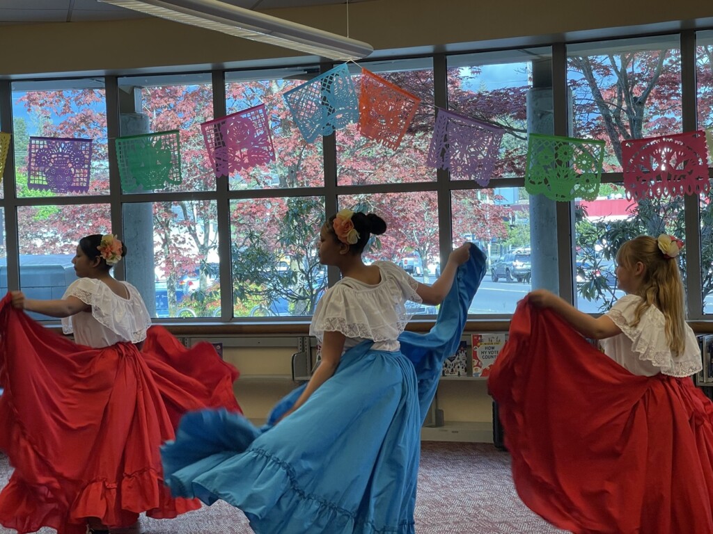 Three young women wearing traditional baile folklórico dress, with white blouses and red or blue skirts and large flowers in their hair hold their skirts out during a dance move in a sunlit classroom.