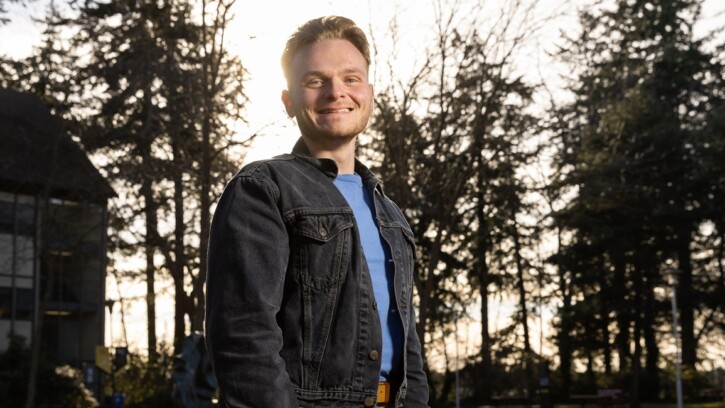 Gavin Ripka, wearing a blue t-shirt and grey denim jacket, stands outside with the sun and large evergreen trees behind him on campus