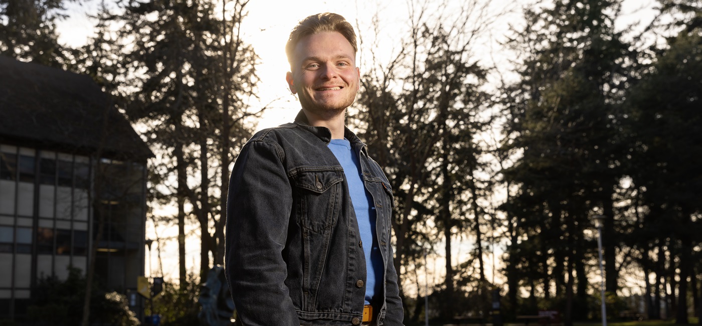 Gavin Ripka, wearing a blue t-shirt and grey denim jacket, stands outside with the sun and large evergreen trees behind him on campus