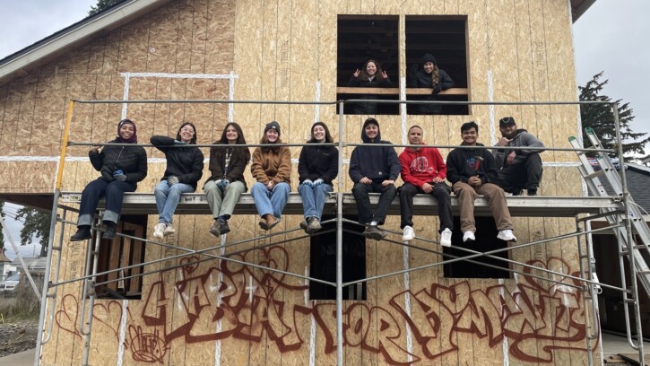 A group of students from the J-Term on the Hill Class sit on scaffolding against a house being built with Habitat for Humanity. Below is graffiti on the unpainted house that reads 'Habitat for Humanity'.