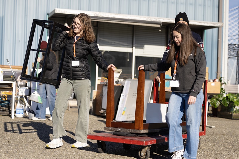 Students outside in a parking help on a sunny winter day push a cart with furniture on it while helping sort and organize furnishings at Northwest Furniture Bank.