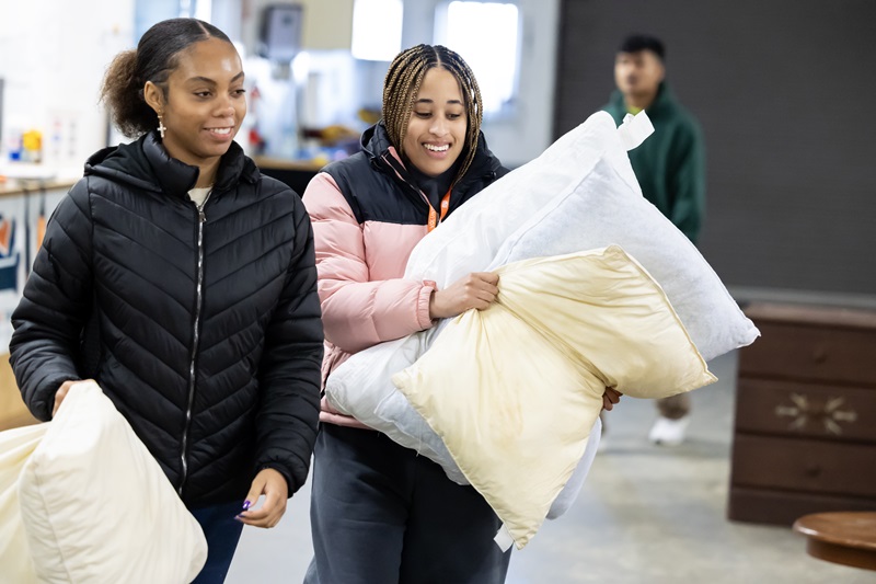 Two students carry pillows inside a warehouse while helping sort and organize furnishings at Northwest Furniture Bank.