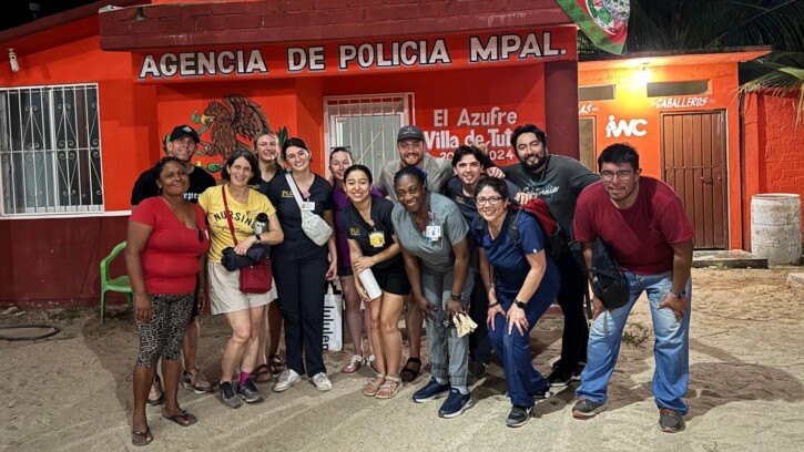 A group of PLU Nursing students and faculty stand together for a posed photo in front of a small red building that reads in white letters above the door "Agencia de Policia MPAL"