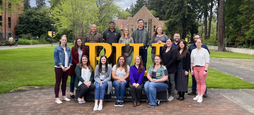 A group of principals surrounding the block letters PLU on campus with buildings and trees in the background.