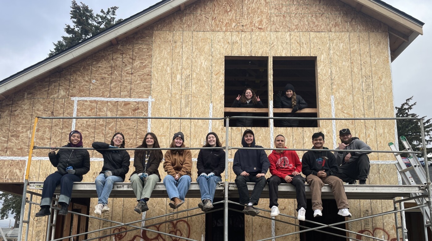 Several students sitting on a scaffold outside of a newly constructed house