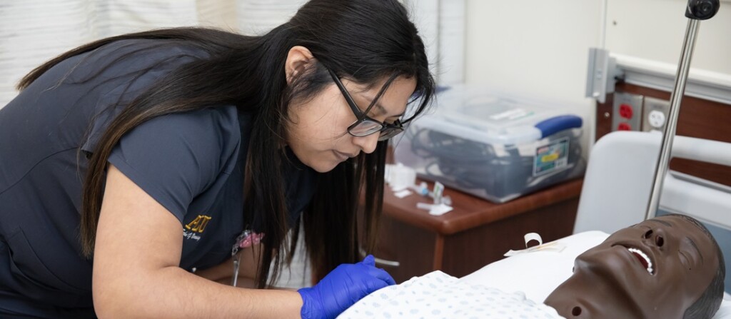 A PLU Nursing student wearing scrubs and gloves bends over a hospital bed and simulated patient in PLU's Nursing Simulation Center