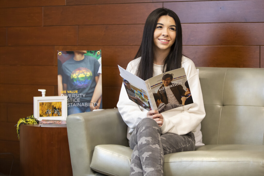 Student holds a brochure while sitting on a couch and smiles into the camera.
