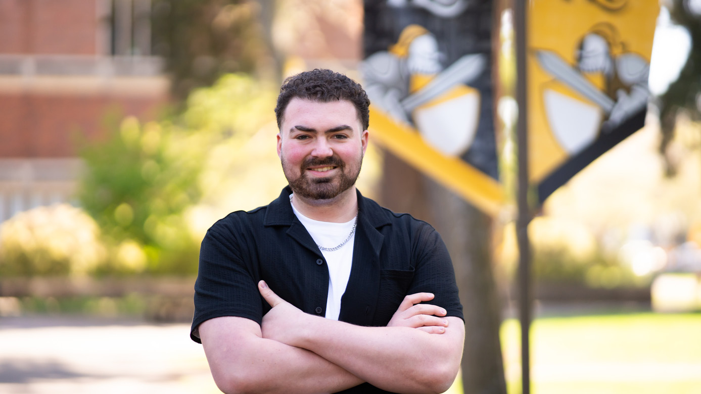 Kaden Bolton smiles while standing with his arms crosssed. He's standing on PLU's cmpus with two black and gold PLU flag pole banners behind him.