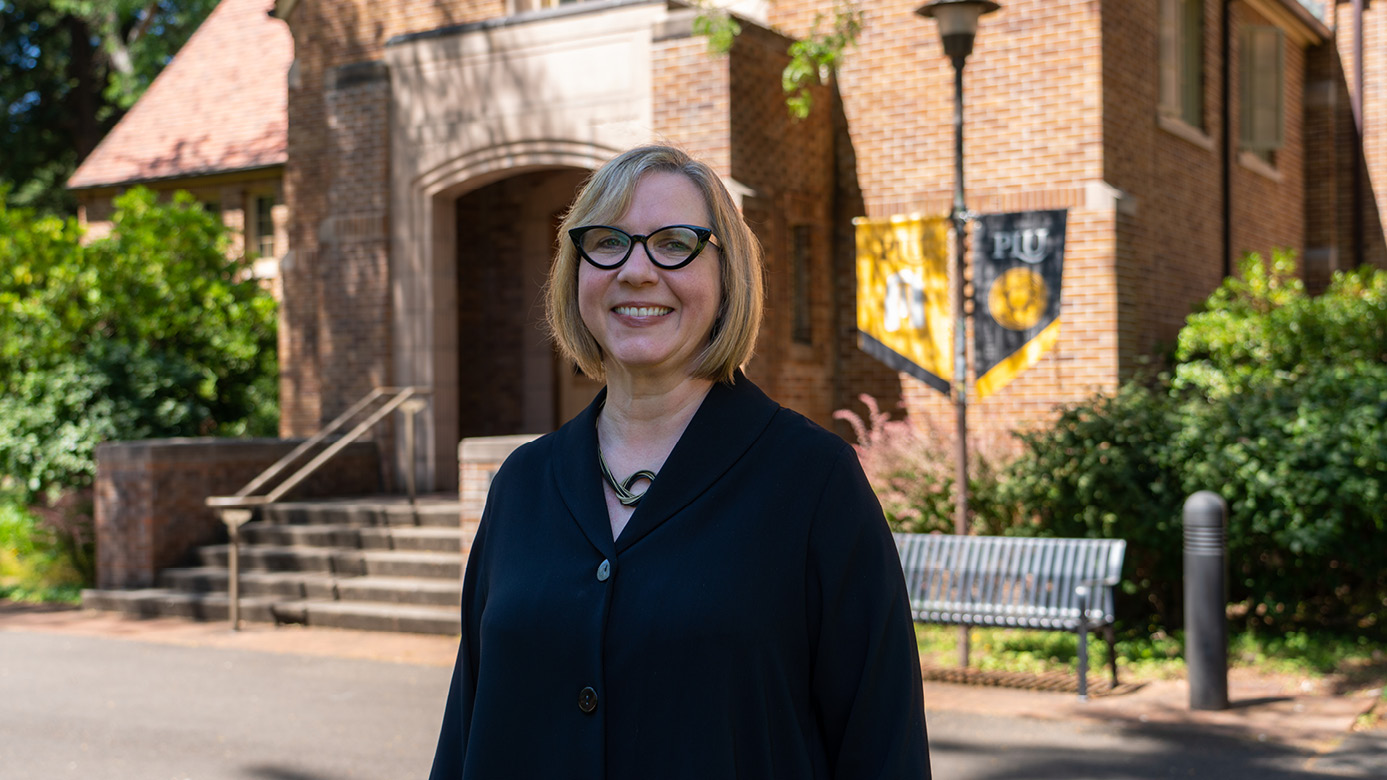 Stephanie Johnson standing outside of PLU's Xavier hall. She's wearing a black blouse and black reading glasses. She's smiling and a gold and black PLU-brand banner is hanging from a light pole in the background.