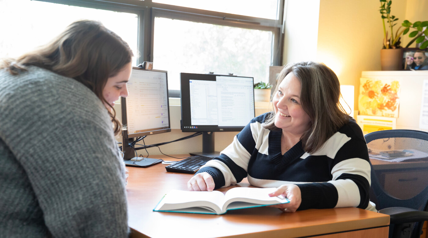 A professor sits at a desk and looks at a book with a student