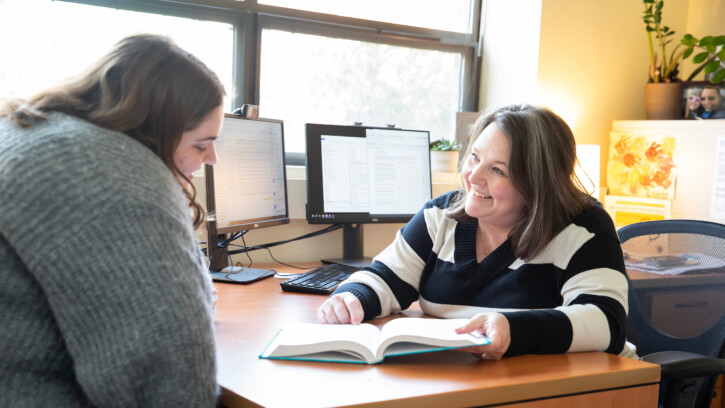 A professor sits at a desk and looks at a book with a student