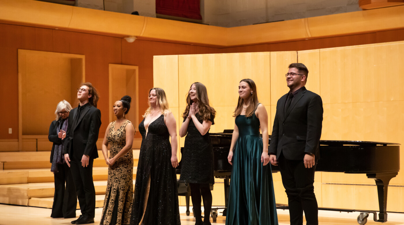 Students stand at the front of the stage in front of the piano