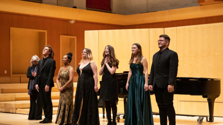 Students stand at the front of the stage in front of the piano