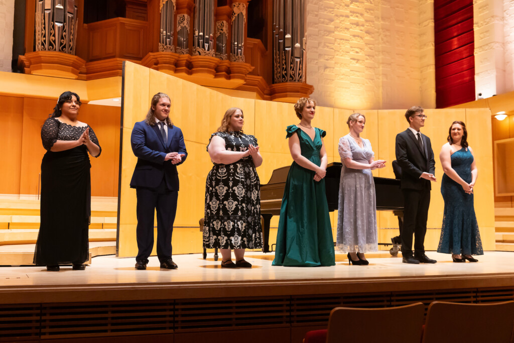 Seven finalists stand on a stage in front of a piano.