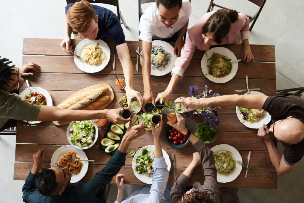 8 people sit at a wooden rectangular table. They are all eating salad or noodles and are clinking their glasses.